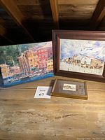 Three framed seaside artworks: two large paintings and one small framed print, displayed on wooden surface.
