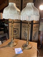 Pair of tall gold-tone candle stick lamps with off-white fringe trimmed lamp shades, set on wooden surface with a screen backdrop.