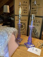 Pair of vintage purple acrylic candlestick table lamps on a wooden surface with visible cords and bulbs installed, showing crystal-like translucent material and square bases.