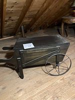Full view of small dark wooden garden cart with metal wheels placed on wooden floor under angled roof beams.