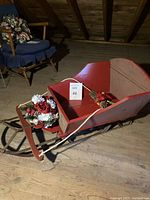 Full side view of the vintage wooden sled showing the red-painted baby seat, box attachment, and floral decoration on the front. The sled rests on a wooden floor.
