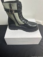 Side view of one black and gray lace-up boot with buckle, on top of white box