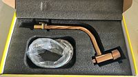 Faucet with curved spout, hose, and mounting hardware inside foam-padded yellow box, sealed with plastic wrap on the hose, indicating new condition.