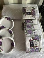 Bundles of white tissue paper, stacks of small floral purple and white paper plates, and floral napkins with purple flowers and green stems shown together.