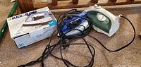 Two electric irons on a carpeted floor with tangled cords, one with its original box labeled Black & Decker Travel Iron.