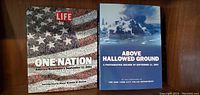 Photo of two large hardcover coffee table books lying flat on a brown wooden surface. Left book titled 'Life One Nation: America Remembers September 11, 2001' with a cover showing American flag made of photographs. Right book titled 'Above Hallowed Ground: A Photographic Record of September 11, 2001' featuring a smoky image of the World Trade Center attacks.