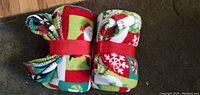 Two wrapped fleece blankets with Christmas themed prints, folded and secured with red straps, placed on carpeted floor.
