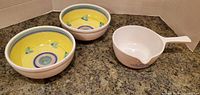 Photo showing two yellow ceramic serving bowls with floral and circular patterns and one white ceramic handled bowl with text on the side arranged on a dark countertop.