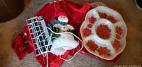 Photo showing red Christmas tree skirt, white snowman figurine wearing a hat and scarf, white string lights, and a serving tray featuring red poinsettia designs.