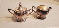 Front view of the silver-tone lidded sugar bowl and creamer showing reflective metal surfaces, ornate handles, and pedestal bases.