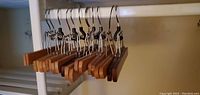 Photo showing nine men's wooden pants hangers hanging from a metal closet rod. Each hanger has a wooden bar with two adjustable metal clips and a metal hook for hanging.