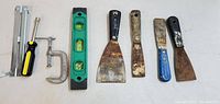 Photo of nine hand tools arranged horizontally showing various spackle knives, a spirit level, clamp, screwdriver, and a sliding T-bevel.