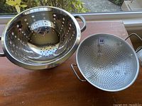 Photo showing two colanders, a stainless steel colander with polished finish and an aluminum colander with matte finish, both on a wooden surface.