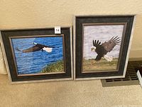 Two framed photographs of bald eagles, one in flight over water, the other on ground with wings spread, both square frames approx 13x12 inches.