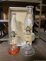 Photo shows two vintage glass oil lamps, one with an orange textured glass base and the other with a clear textured glass base. Both have clear chimneys and metal burners.