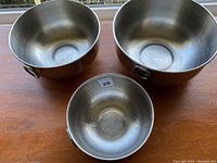 Three stainless steel bowls of varying sizes shown on wooden surface. Two have side handles, one does not. Appears lightly used.