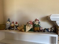 Eight painted garden gnome figurines displayed on a shelf, showing variety in sizes, poses, and colors including blue, red, green hats, and detailed clothing.