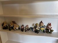 Front view of nine assorted garden gnome figurines arranged on a white shelf, showing varying sizes and poses with colorful painted details.