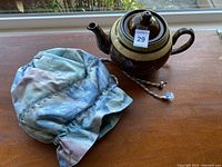 Brown ceramic teapot with light beige band and matching lid, alongside blue-purple tie-dye fabric tea cozy with drawstring, placed on wooden surface by a window.