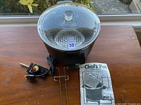 Electric Chef's Pot with glass lid, metal basket and heating element with cord on wooden table near window
