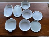 Nine pieces laid out on wood surface: 3 round dishes, 2 round lids, 1 square dish with handles, 1 oval dish, 1 oval lid