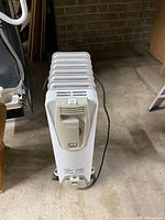 Photo of white Delonghi oil heater with multiple fins, power cord seen, standing on floor against brick wall, untested.