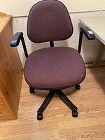 Front view of mauve office chair showing padded seat, backrest, black armrests, and rolling casters on wooden floor near wooden and plastic furniture.