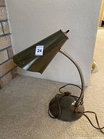 Full side view of a dark metal desk lamp with an adjustable gooseneck arm and perforated conical shade sitting on carpet in front of a brick wall.