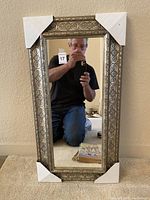Full view of the decorative rectangular mirror with a metallic ornate frame and corner protectors on all four corners. The mirror is reflecting the person taking the photo.
