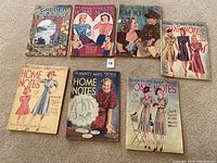 Top view of 7 vintage magazines laid on carpet: 6 Home Notes and 1 Garden Beautiful from 1940s with illustrated covers depicting fashion and gardening.