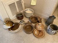 Photo of various pottery pieces arranged on carpeted floor including bowls, jars, pitchers, and covered dishes in diverse glazes and styles.