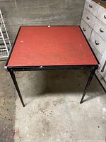 Front angled view of red square card table with black metal legs and frame, some marks on table surface.