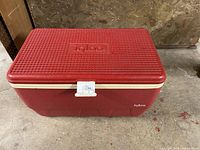 Red rectangular Igloo cooler with textured lid and embossed Igloo logo, white trim and latch, sitting on concrete floor against plywood wall.