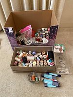 Two boxes on floor containing numerous spools of sewing thread, packaged needles, bobbins, bobbin cases, and small sewing accessories.