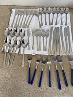 Array of stainless steel utensils arranged on a white cloth, including multiple forks, spoons, knives, and additional forks with blue handles at the bottom