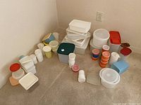 Various assorted Tupperware plastic containers with lids arranged on a carpet floor in a corner of a room.