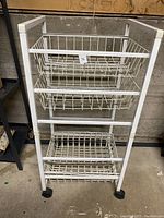 White metal wire shelving rack with three wire basket shelves, standing on a concrete floor, against a wall.