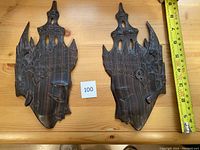 Pair of dark wooden carved plaques showing stylized faces with tall headdresses, close to a measuring tape showing height approx. 16 cm.