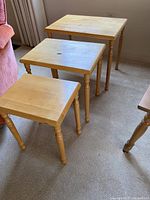 Three solid wood nesting tables arranged in ascending size, showing square tops and turned legs, placed on carpet near sofa and curtain.