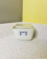 Clear front view of white opaque Pyrex fridge dish with blue snowflake decoration and fitted clear glass lid, showing minor discoloration.
