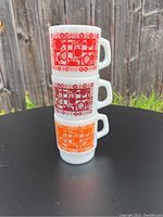 Three stacked white milk glass mugs with red and orange fruit block patterns, stacked vertically against a wooden fence surface.
