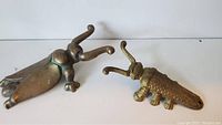 Two beetle-shaped shoehorns, one brass and one darker metal, placed on a white surface.