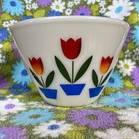Side view of FireKing large white glass bowl with colorful tulip floral pattern on exterior, sitting on a multi-colored floral fabric background.