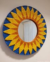 Photo of a round wall mirror with textured wooden frame painted in blue, yellow, and orange sunburst design, hanging on a wall.