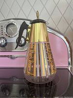 Front angle view of the Fred Press carafe showing geometric gold design, brass top, black handle, and finial.