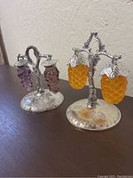 Grape-themed salt and pepper shaker sets with detailed silver-tone bases and holders, photographed on a wooden surface with a white textured wall background.
