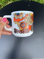 Mug held in hand showing bright retro floral and geometric patterns in orange, yellow, black and white on opaque glass.