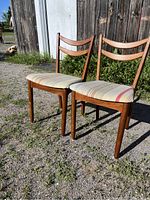 Two mid century walnut dining chairs outdoors with wooden double slat backs and striped fabric seats, side and front angle view.