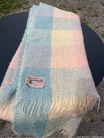 Pastel mohair blanket folded with visible checkered blocks in pastel blue, pink, cream, and yellow, placed on a black surface outdoors.