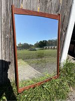 Full view of the mid century teak framed mirror leaning against a weathered wooden wall outdoors.